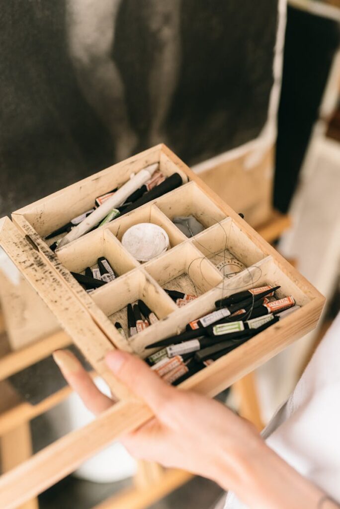 A hand holding a box of various art supplies in a creative studio setting.